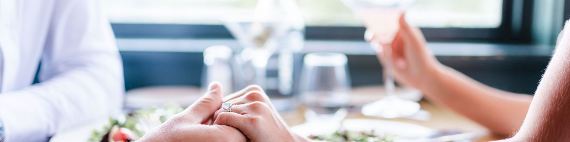 Two individuals are sitting at a table, holding hands, with a salad and drinks in the background.