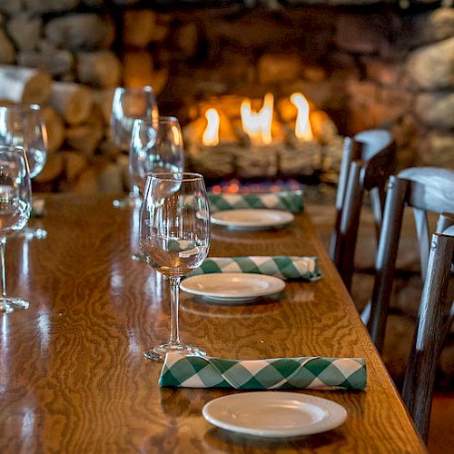 A cozy dining table setting with wine glasses, plates, and green checkered napkins is ready near a stone fireplace.