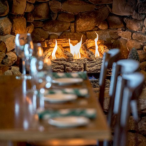 A cozy dining setting with a wooden table, wine glasses, and a stone fireplace with a blazing fire in the background.