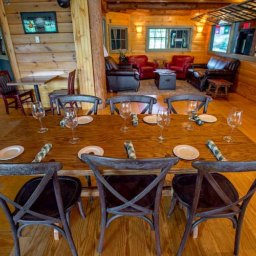 This image shows a cozy restaurant interior with a wooden table set for six, featuring wine glasses and plates, with a lounge area in the background.