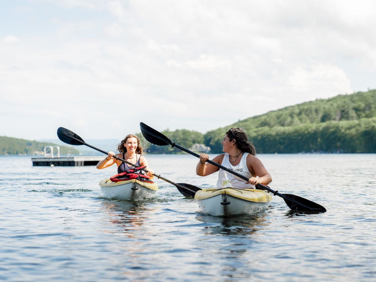 Two people are kayaking on a calm body of water with a dock in the background, surrounded by lush green hills under a partly cloudy sky.