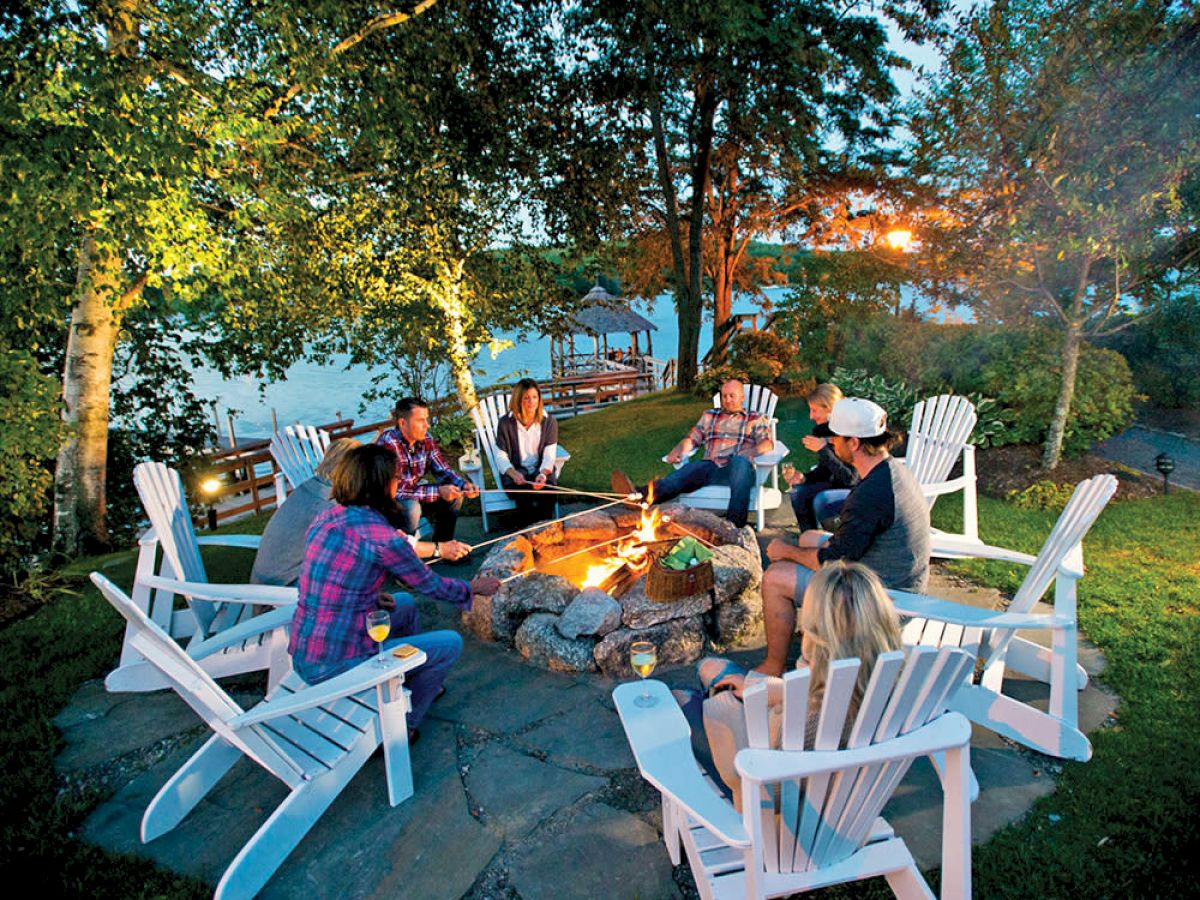 A group of people sits in a circle of Adirondack chairs around a fire pit, surrounded by trees, with a body of water in the background ending the sentence.
