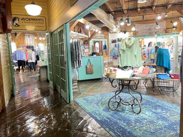 This image shows the interior of a clothing store named Adornments, featuring racks of clothes, a blue rug, a glass table, and wooden decor.