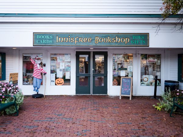A storefront with a sign that reads "Innisfree Bookshop," featuring books, cards, toys, and gifts. Decor includes a scarecrow and a chalkboard sign.