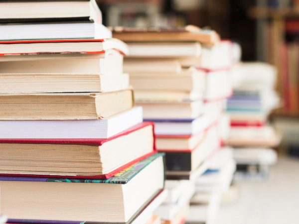 The image shows multiple stacks of books on a table or surface, with more bookshelves visible in the blurry background.
