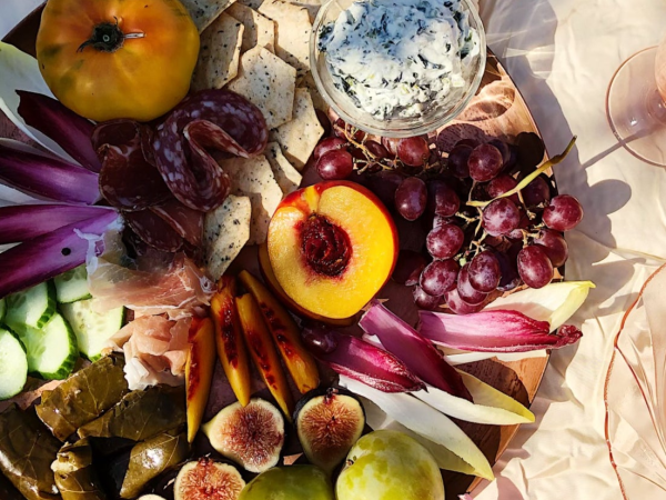 A wooden platter with cheese dip, grapes, peach, figs, crackers, cured meats, radicchio, and cucumbers on a floral tablecloth.