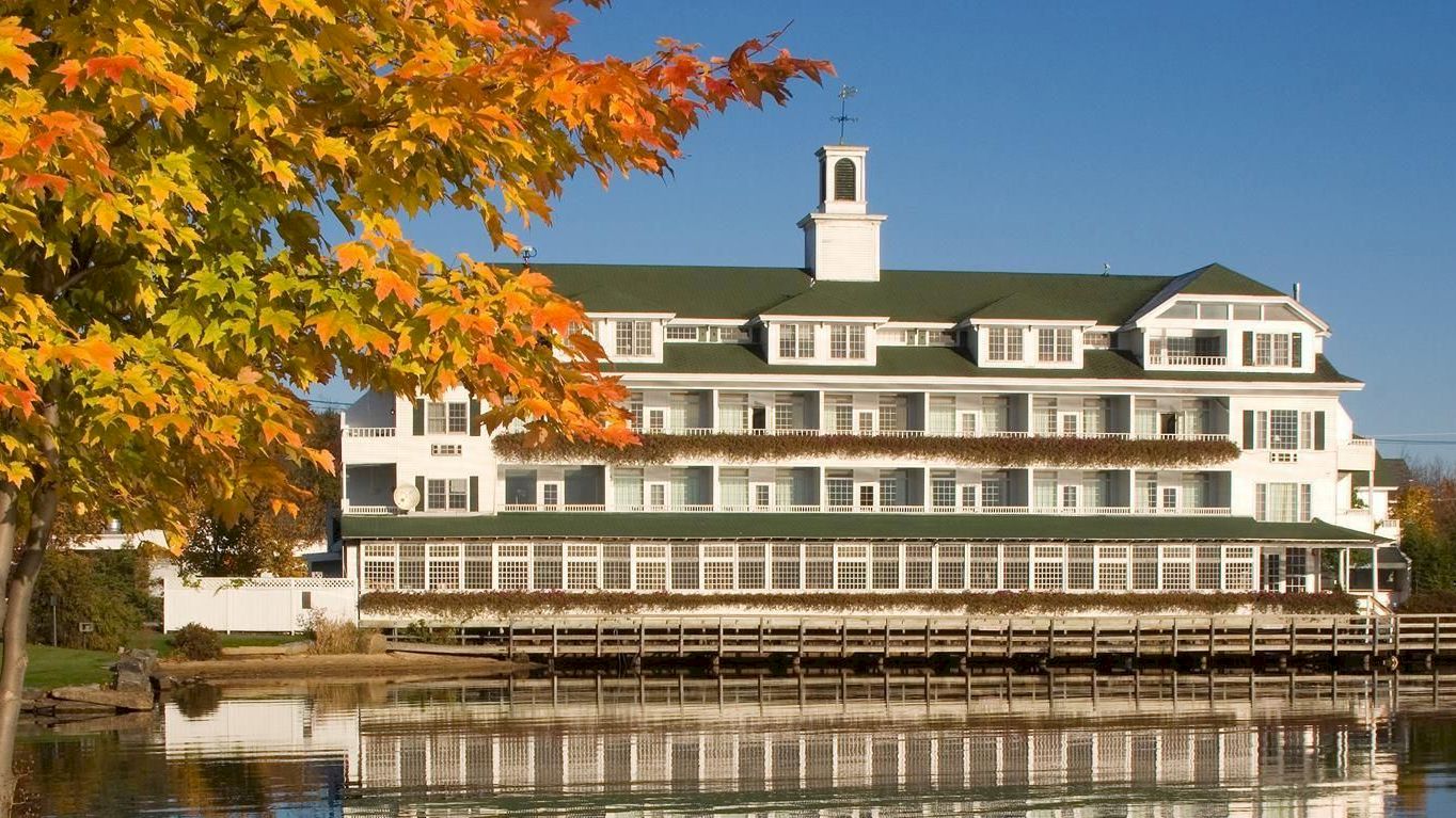 This image shows a large building by a lakeside with vibrant autumn foliage and clear blue skies, reflected in the calm water.
