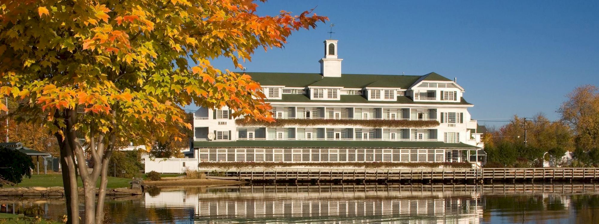 This image shows a large building by a lakeside with vibrant autumn foliage and clear blue skies, reflected in the calm water.
