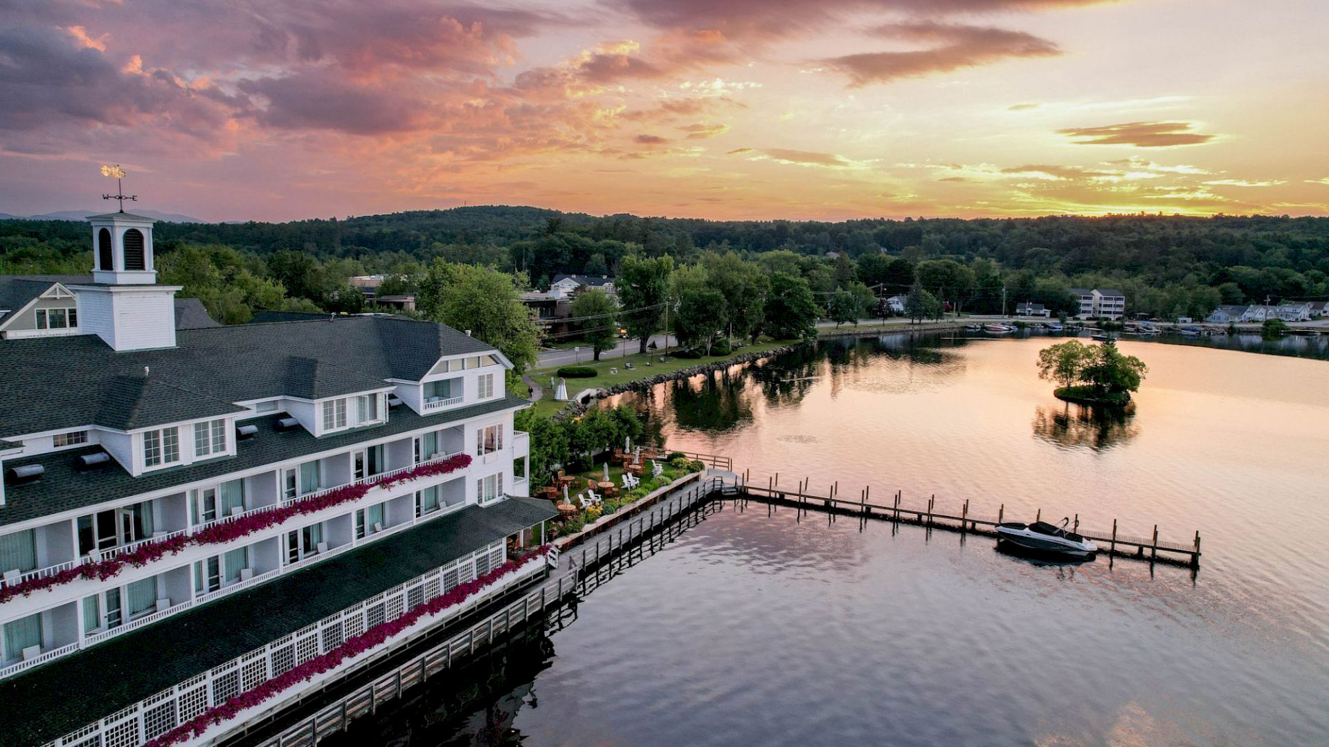 A beautiful lakeside building with a dock extending into the water during a picturesque sunset, surrounded by trees and a small island.