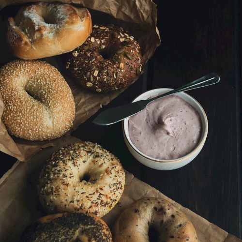 A spread of assorted bagels (plain, sesame, poppy, sesame-seed crust, and whole-grain) with a pink-tinted cream or yogurt dip on the side, all on brown parchment.
