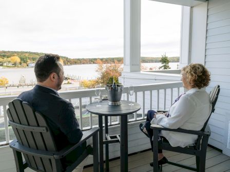 Two people sit on a balcony with a small table and potted plant, enjoying a lakeside view in a calm, sunny setting.