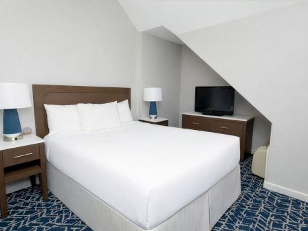 A hotel room with a double bed, two nightstands with lamps, a dresser with a TV, and a sloped ceiling in a blue patterned carpet.