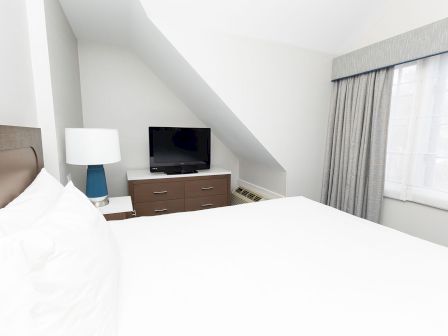 A bright hotel room with a large bed, a dresser, a flat-screen TV, a blue lamp, and a window with light curtains.