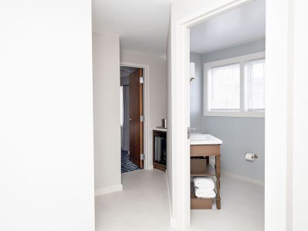 A bright bathroom and hallway: white walls, a wooden vanity with towel shelf, a windowed corner, and an open doorway leading to another room.