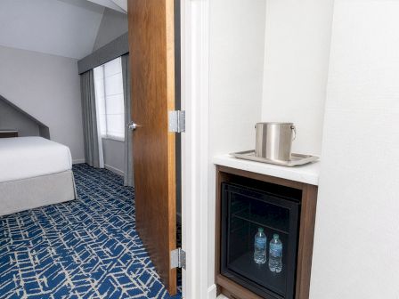 A hotel room with a bed, blue patterned carpet, and an open door showing a small cabinet with a mini-fridge and water bottles; a desk area is visible in the background.