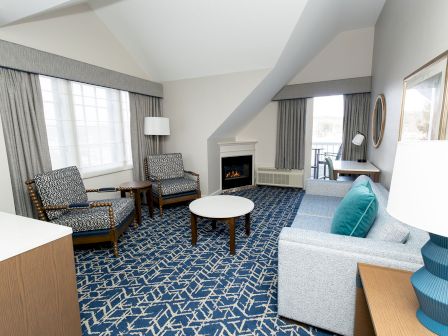 A hotel living room with a blue patterned carpet, seating area, fireplace, large windows, and a corner desk, all under a sloped ceiling.