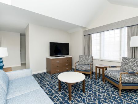 A bright hotel living area with a blue patterned carpet, light furniture, two armchairs, a sofa, a round coffee table, a small TV on a wooden dresser, and large windows.