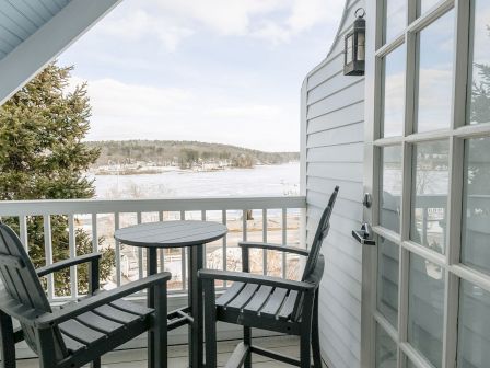 A cozy balcony with a small round table and four dark chairs, overlooking a snowy lake and distant hills, near a white house with glass doors.