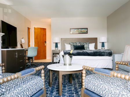A modern hotel suite with a king bed, blue accents, a seating area, a round coffee table, and a desk, bathed in soft lighting.