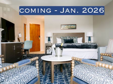 The image shows a hotel room with a modern design, featuring a bed, chairs, a TV, and a desk. Text reads "COMING - JAN. 2026".