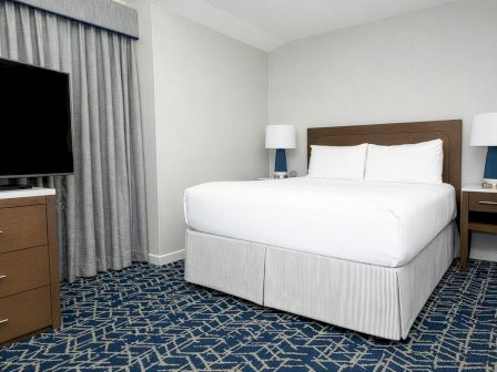A hotel room with a white bed, two nightstands with lamps, a dresser, a flat-screen TV, and blue patterned carpeting, neat and simple.