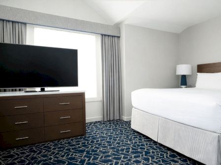 A modern hotel room with a large bed, a dresser with a flat-screen TV, a window with curtains, a lamp, and a patterned blue carpet.