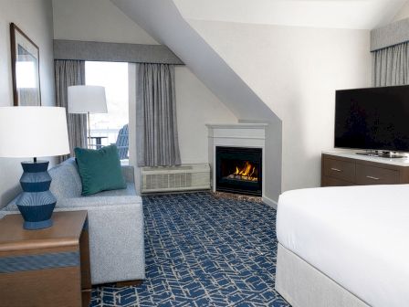 A cozy hotel suite with a sofa, blue patterned carpet, desk lamps, a fireplace, and a flat-screen TV on a dresser by the window.