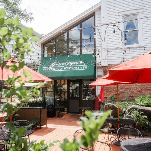 Outdoor cafe scene with green awning, string lights, red umbrellas, and a wooden deck in front of a building. A cozy dining spot.