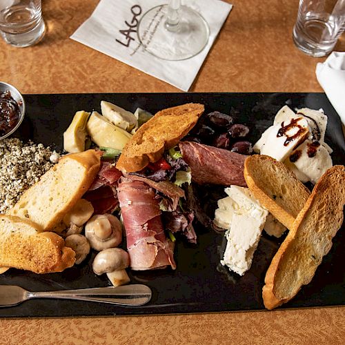A charcuterie board with sliced meats, cheeses, bread, mushrooms, and a jam, served on a black slate with utensils and napkin in the background.