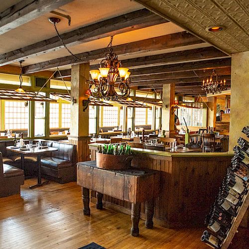 The image shows a cozy, rustic restaurant interior with wooden beams, chandeliers, cushioned booths, tables, and a wine rack set beside a wooden bar.