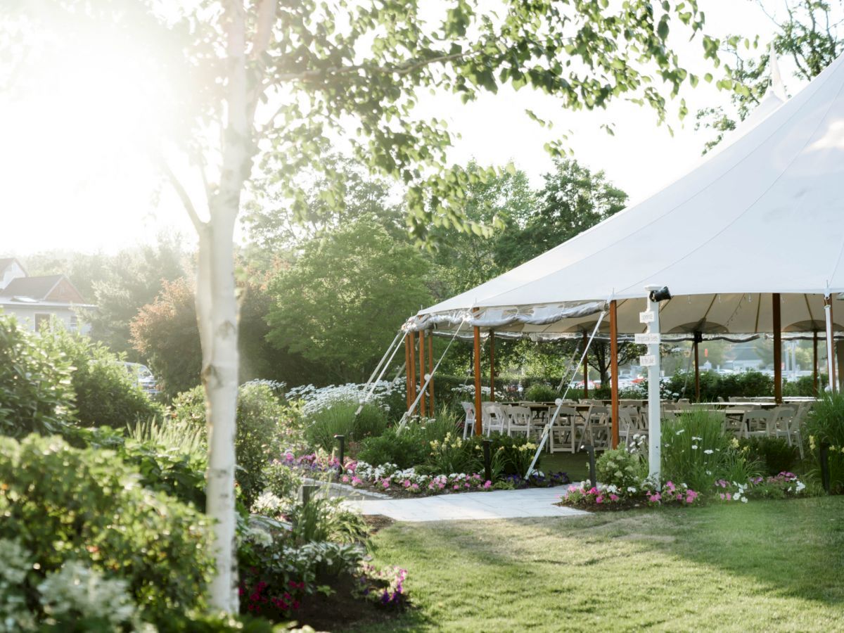 A scenic garden with a white tented event area surrounded by greenery and colorful flowers, trees, and a pathway leading to the tent.