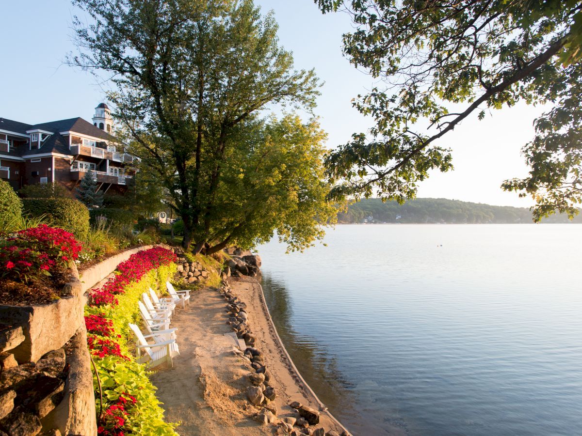 A lake with a sandy shore, white chairs, a tree, a building with balconies, and flowering plants under a clear sky at sunset.