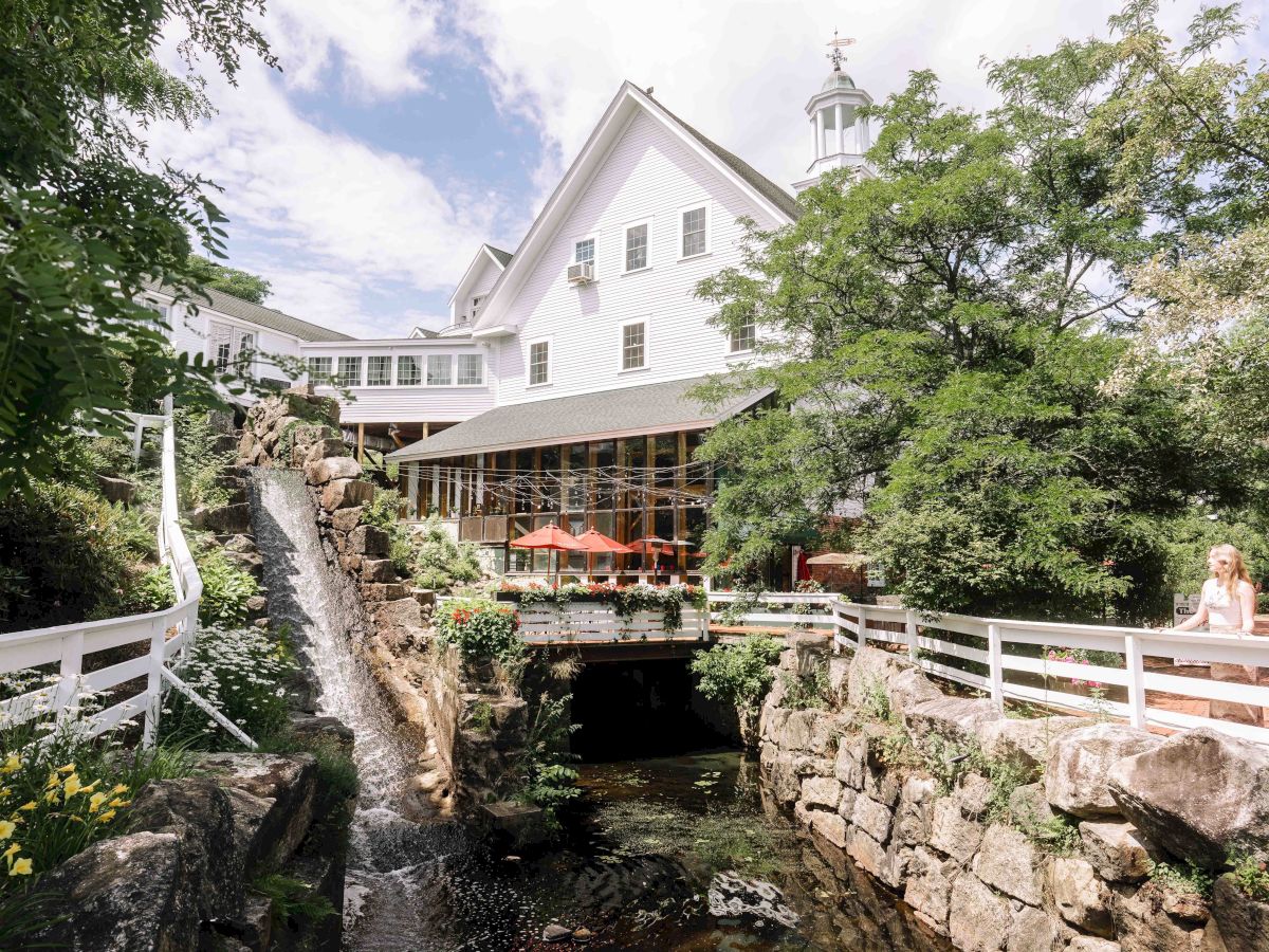 The image shows a picturesque building with a white facade surrounded by lush greenery, a waterfall, outdoor seating, and a small bridge.