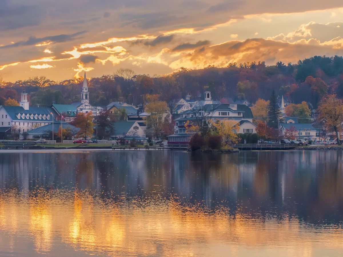 A picturesque town by a lake during sunset, with autumn foliage, reflections on the water, and a serene, peaceful atmosphere ending the sentence.