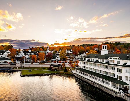 A scenic view of a waterfront town during sunset, with autumn-colored trees, a white building along the water, and charming houses in the background.