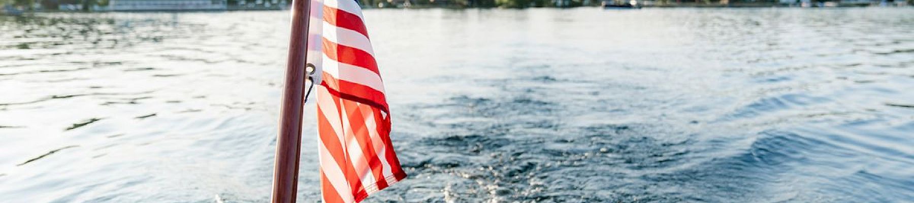 A person is boating on a lake, holding the edge with a flag pole displaying the American flag at the back of the boat.