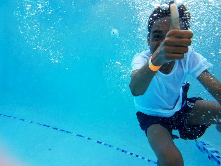 A person underwater in a pool giving a thumbs up, wearing a white shirt and blue shorts, with bubbles and lane lines visible.