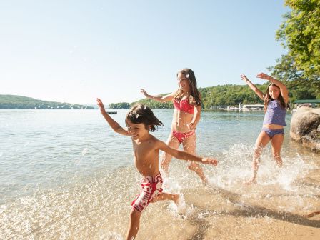 Kids having fun at the beach, running along the shore as waves splash around them, sunny day, clear sky, and trees in the background.