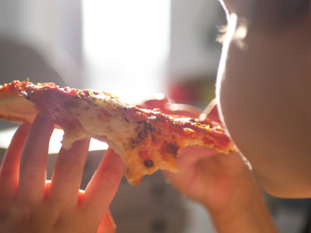 A person is enjoying a slice of pizza, holding it up to their mouth and taking a bite, with the backlit image capturing the pizza details.