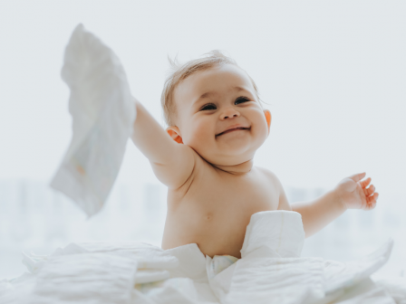 A cheerful baby is sitting and playing with diapers, holding one up in the air while surrounded by several others.