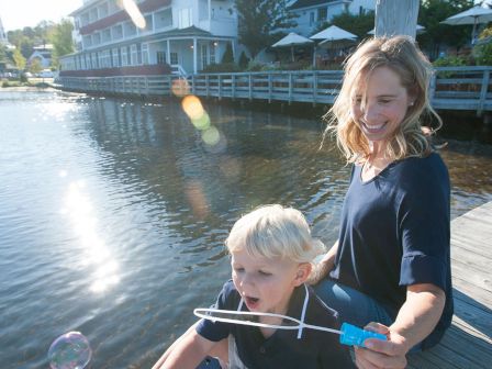 Two children on a wooden dock by the water, blowing bubbles with a bubble wand, a large house and blue sky in the background.