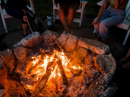 Three individuals are seated around a campfire, roasting marshmallows on sticks while the fire burns brightly between them.