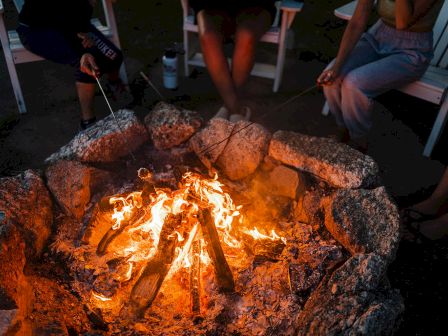 People gathered around a campfire roasting marshmallows or snacks, seated on chairs in a circle.