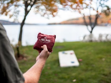 A person holds a bean bag, playing cornhole outdoors near a lake, with trees in the background and a cornhole board in the distance.