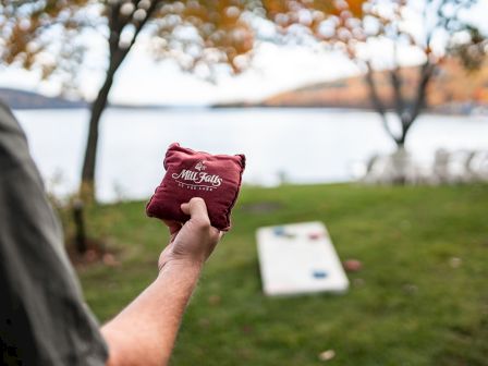 A person holding a sandbag playing cornhole with a lake, trees, and a white board on a grassy shoreline background.