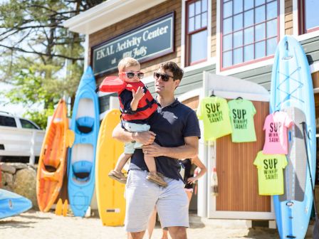A man holding a child near kayaks and paddleboards at an activity center, with colorful t-shirts that read "SUP in the USA."
