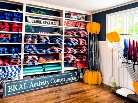 A neat storage area with labeled shelves full of colorful dry bags, life vests, and gear, plus paddles and canoes/kayak equipment near a window, EKAL Activity Center kayak gear.