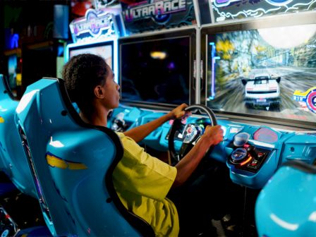 A person is playing a racing arcade game in a blue arcade cabinet, focused on a car chase on the screen, in a dim arcade.