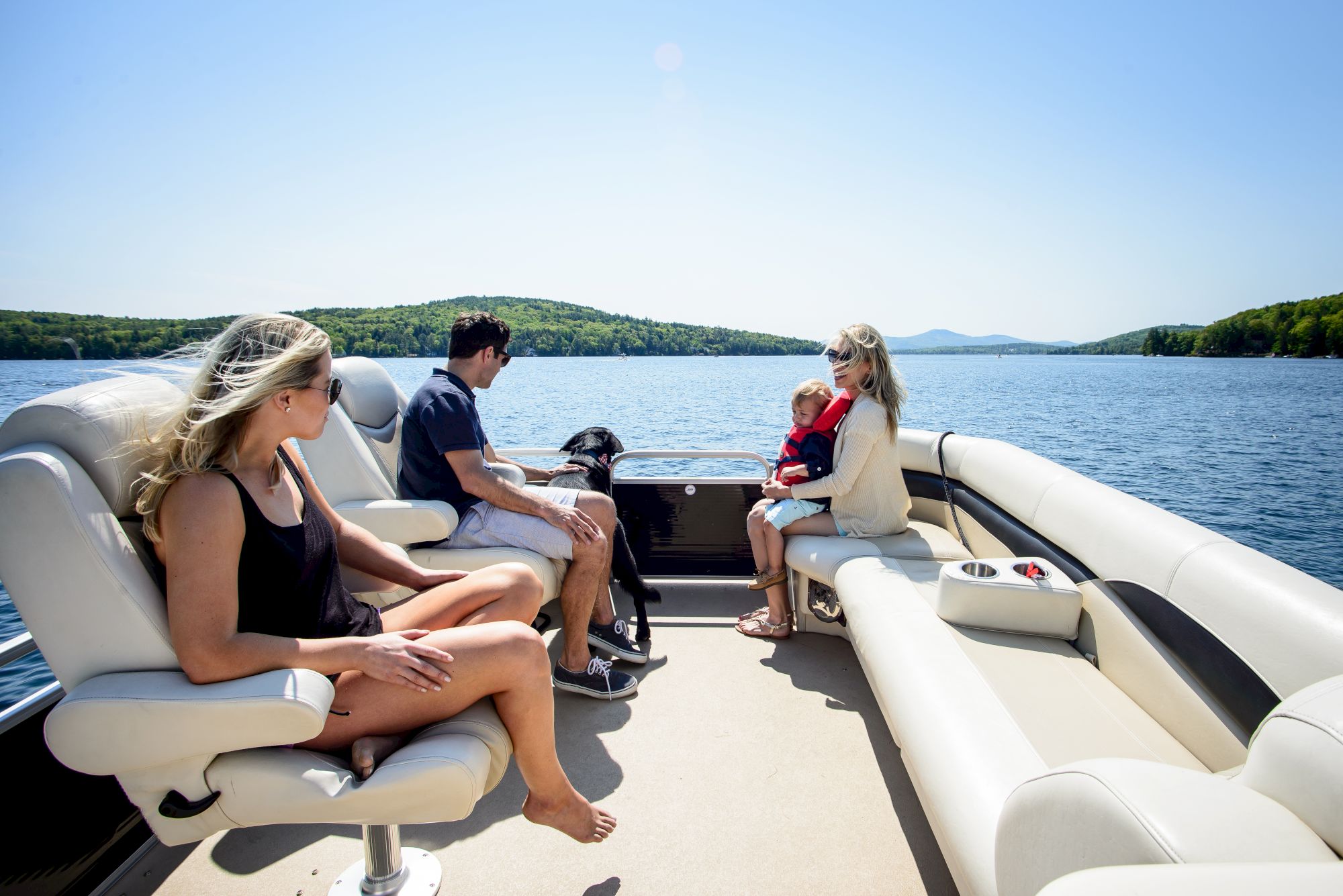 Four people and a dog are on a boat on a sunny day, with trees and hills in the background.