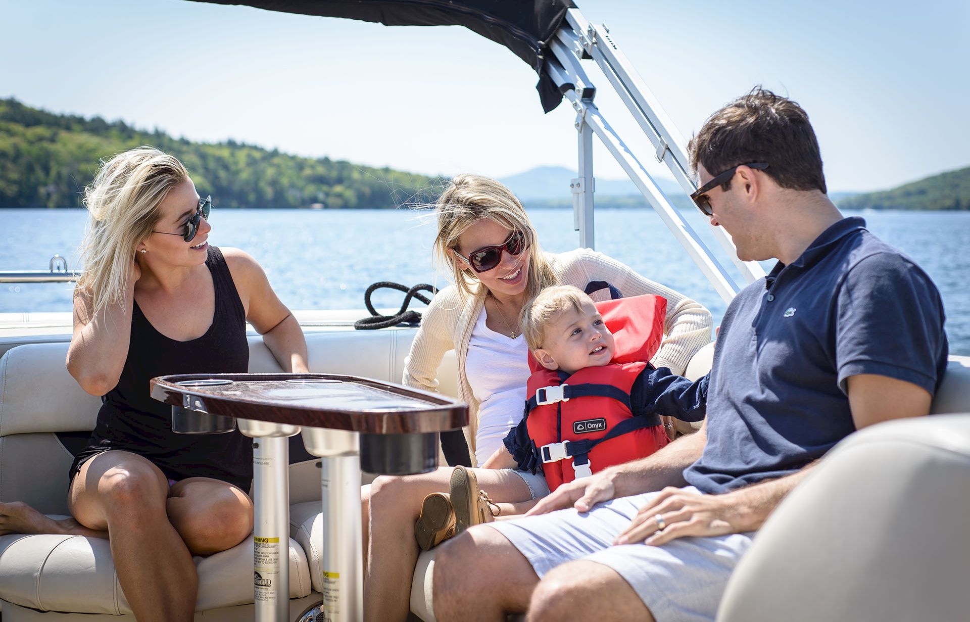 A family on a boat: two women, a man, and a small child in a life jacket, sitting around a table with water and hills in the background.